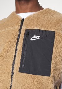 Beige fleecetröja med dragkedja fram, med en svart dragkedjeficka och en vit Nike-logotyp på fickan. Mjuk textur med en avslappnad passform.