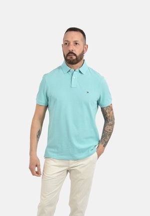 Mann mit kurzem Haar und Bart, der ein hellblaues Poloshirt und beige Hose trägt, mit einem floralen Tattoo-Ärmel am rechten Arm.