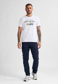 Bílý tričko s zeleným grafickým textem "PETROL IND. EST. 1989," v kombinaci s námořnickými cargo kalhotami a šedými teniskami.