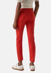 Rote, schmal geschnittene Baumwollhose mit Gesäßtaschen und einem Bundetikett. Kombiniert mit weißen Sneakern mit brauner Sohle.