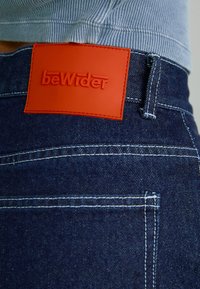 Mørke jeans i denim med kontrasterende hvit søm, som har et teksturert rødt rektangulært lærmerke merket "beWider" på linningen.