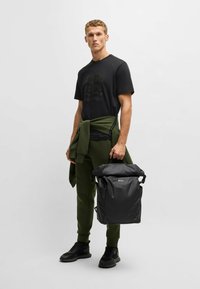 Sort roll-top rygsæk med logo, to sidelommer og justerbare stropper. Båret af en model i olivengrønne joggers og sorte sneakers.