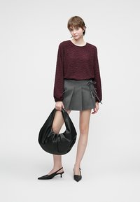 Top a maniche lunghe di tessuto bordeaux abbinato a una gonna grigia a pieghe. Porta una grande borsa in pelle nera. Scarpa nera a punta. Sfondo bianco.