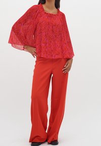 Blouse rouge plissée avec un design évasé et un col froncé, associée à un pantalon rouge à jambes larges. La blouse présente un motif floral dans des nuances de rouge et de rose.