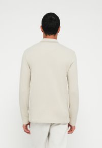 Polo de manga comprida em bege claro com acabamento texturizado, gola e bainha reta, combinado com calças de cor clara.