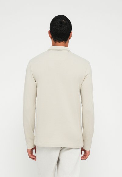 Polo à manches longues beige clair avec une finition texturée, col et ourlet droit, associé à un pantalon de couleur claire.