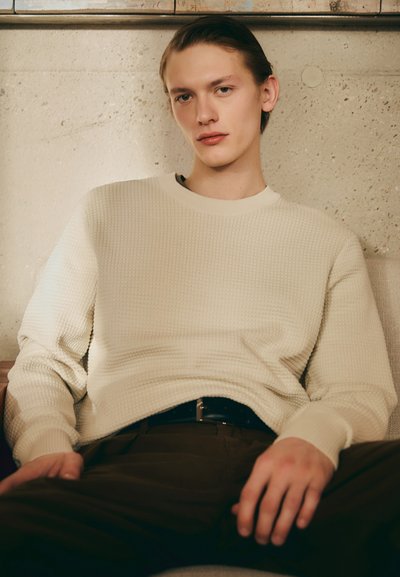 Lys beige tekstureret sweater med ribbet mønster, rund hals og lange ærmer, kombineret med mørke bukser og et sort bælte, siddende op ad en neutral væg.