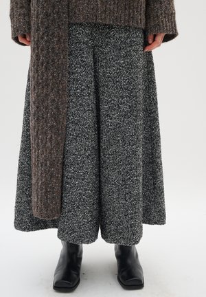 Kobieta ubrana w szarą spódnicę w jodełkę, czarne botki za kostkę i długi brązowy tweedowy płaszcz, stojąca na białym tle.