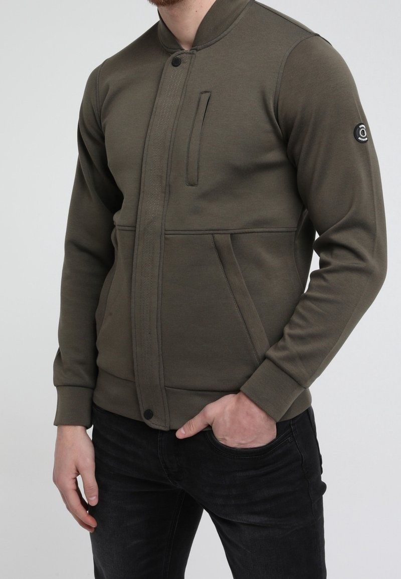 Homme portant une veste bomber zippée vert olive avec poches sur la poitrine et les côtés, et un jean noir, main dans la poche, devant un fond uni.