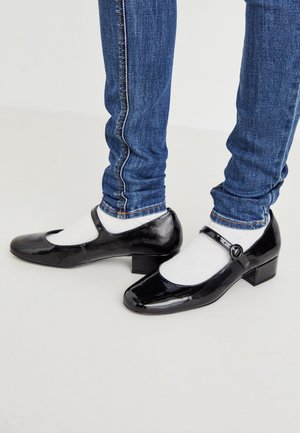 Person wearing blue skinny jeans, white socks, and shiny black Mary Jane shoes with a low heel on a white floor.