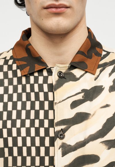 Άνδρας που φοράει πουκάμισο με ανάμεικτα σχέδια: ασπρόμαυρο καρό σε μία πλευρά, μπεζ και μαύρα πινελιές στην άλλη, και καφέ γιακά καμουφλάζ.