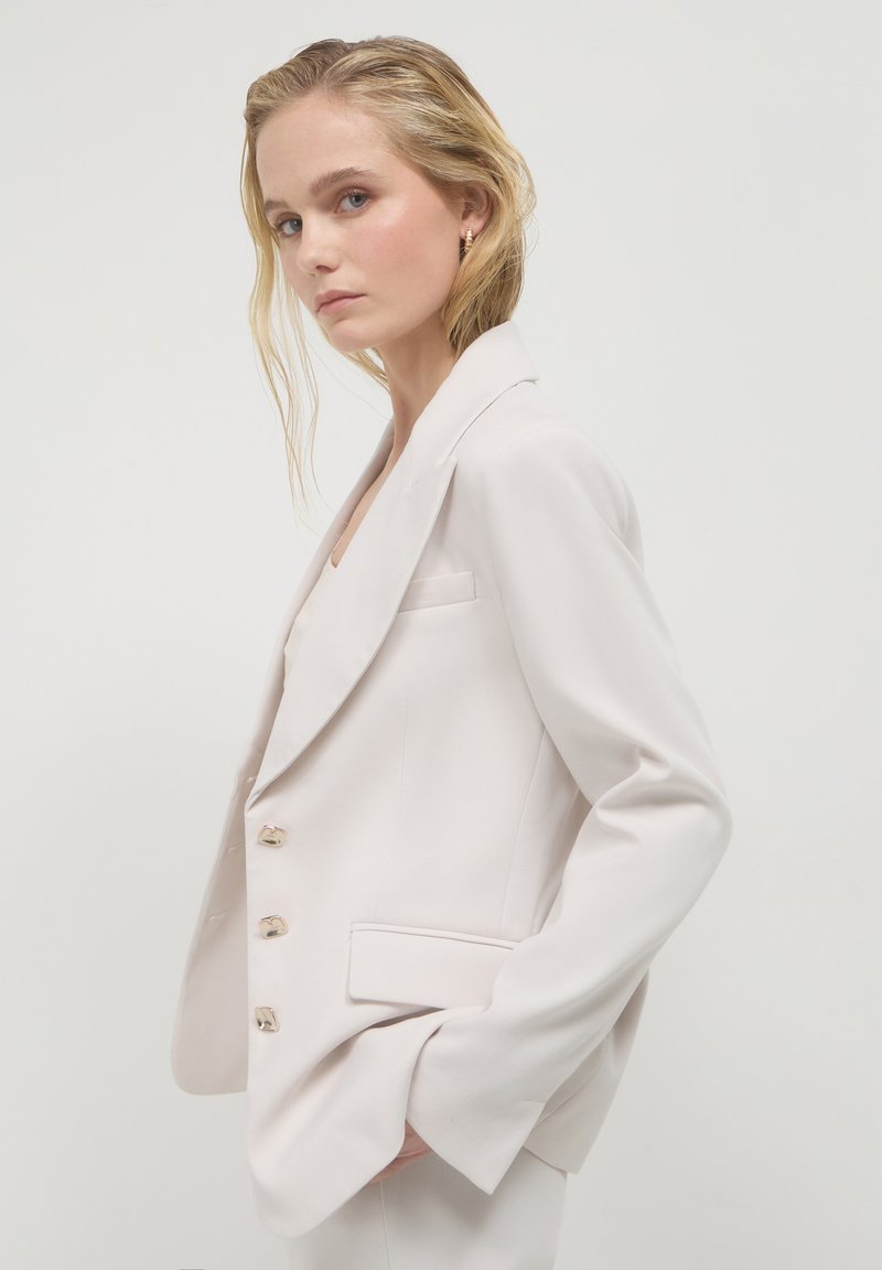 Hellbeige Blazer mit taillierter Passform, aufgesetzten Revers, goldfarbenen Knöpfen und Seitentaschen. Glatte Textur und strukturierte Gestaltung.