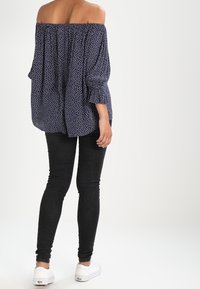 Kvinna i marinblå avskuren blus med vita prickar, svarta tajta jeans och vita sneakers, stående med ryggen mot kameran.