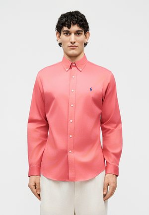 Άνδρας με κοραλί πουκάμισο με κουμπιά και μακριά μανίκια, με μικρό ναυτικό λογότυπο στο αριστερό στήθος, συνδυασμένο με ανοιχτό μπεζ παντελόνι.