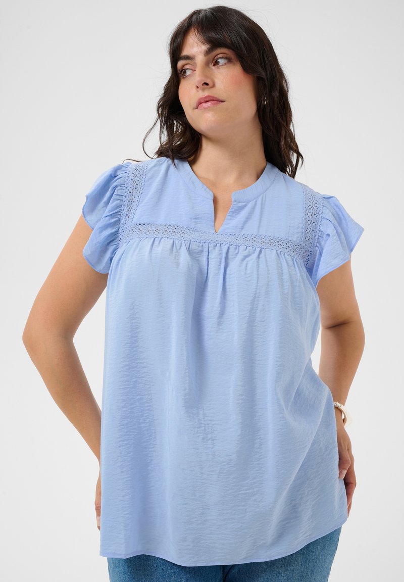 Femme aux cheveux foncés portant un chemisier bleu clair à manches courtes avec un détail en dentelle, debout avec une main sur la hanche, regardant sur le côté.