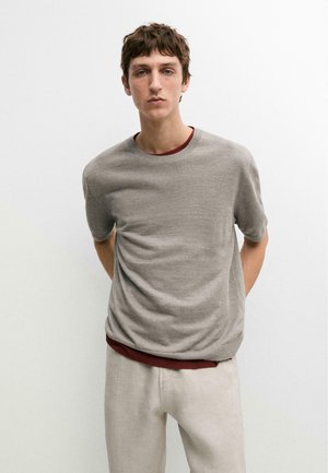 Giovane uomo con capelli castani corti, indossa un maglione grigio a maniche corte sopra una camicia color bordeaux e pantaloni beige chiaro, in piedi contro un muro bianco.