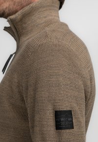 Suéter marrón texturizado con cremallera en el cuello y un cuello acanalado. Presenta un parche de marca negro en la manga, destacando su material y diseño.