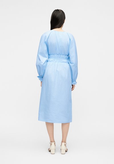 Γυναίκα με μακριά σκούρα μαλλιά, που φοράει ανοιχτό μπλε μεσαίο φόρεμα με φουσκωτά μακριά μανίκια και άσπρα πέδιλα με τακούνι, γυρισμένη πλάτη σε λευκό φόντο.