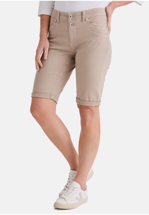 Person trägt beige kniehohe, aufgerollte Shorts, ein weißes langärmliges Oberteil und weiße Turnschuhe und steht mit einer Hand auf der Hüfte.