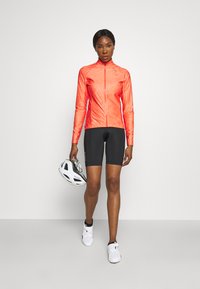 Bright orange cycling jacket with a swirl pattern, paired with black cycling shorts. A white helmet is held in the left hand.