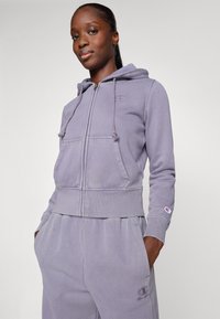 Light purple zip-up hoodie made of soft material, featuring a hood, front pockets, and logo detail on the chest and sleeve.