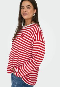 Maglietta a maniche lunghe a righe rosse e bianche con scollatura rotonda e una piccola patch a forma di cuore; abbinata a jeans in denim blu. Vestibilità casual.