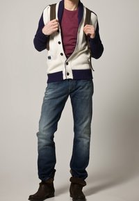 Cardigan blanco y azul marino con botones, camisa marrón, jeans azules y botas de tobillo marrones. Los tirantes de la mochila son visibles en los hombros.