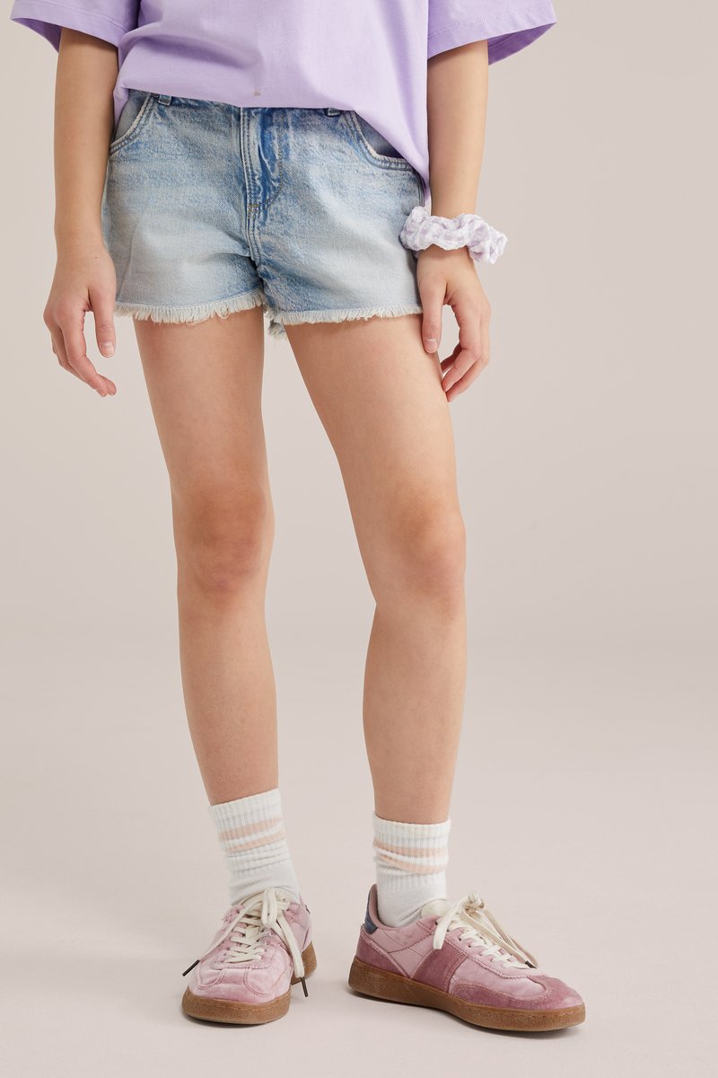 Osoba ubrana w jasne, postrzępione szorty z denimu, lawendową bluzkę, białą frotkę, białe skarpetki z różowymi paskami oraz różowe trampki z brązowymi podeszwami.