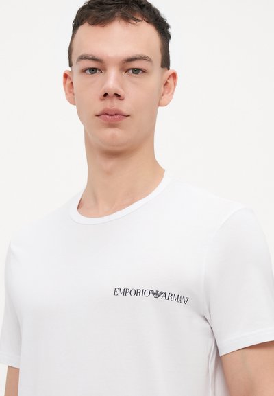 Weißes Baumwoll-T-Shirt mit kurzen Ärmeln, das auf der linken Brustseite ein schwarzes gesticktes "Emporio Armani"-Logo trägt. Glatte Textur.