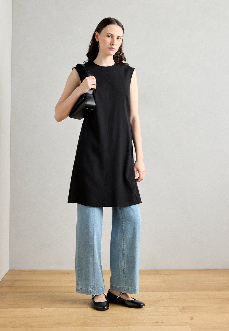 Zwarte mouwloze jurk met een rechte silhouet, gecombineerd met lichtblauwe wijde jeans en zwarte platte schoenen met studaccenten.