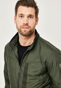 Grüne Zip-Jacke mit hohem Kragen, ausgestattet mit schwarzen Akzenten auf den Schultern und einer glatten Textur. Getragen über einem schwarzen Shirt.