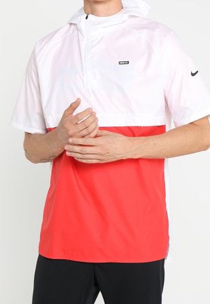 Mężczyzna ubrany w białą i czerwoną bluzę Nike z krótkim rękawem, czarnym suwakiem i czarne spodnie, stoi z rękami złożonymi z przodu.