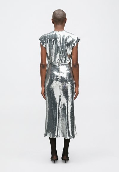 Persona stāv ar muguru pret skatītāju, ģērbusies sudraba pailga kleitā ar spīguļiem bez piedurknēm, melnās zeķbiksēs un melnām augstpapēžu kurpēm uz vienkārša fona.