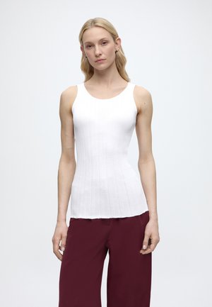 Ξανθιά γυναίκα με λευκό ριγωτό αμάνικο τοπ και μπορντό παντελόνα με φαρδιά μπατζάκια, που στέκεται μπροστά σε απλό ανοιχτόχρωμο φόντο.