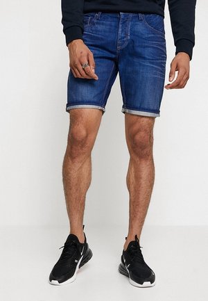 Pantalones cortos de mezclilla azul oscuro con dobladillos enrollados, diseño de cinco bolsillos y cierre de botones. Usados con zapatos deportivos negros.