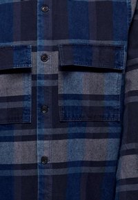 Camisa de cuadros azul con frente abotonado, dos bolsillos en el pecho y distintas tonalidades de azul y gris en una tela texturizada.