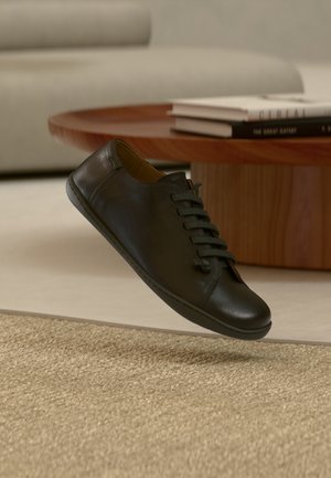 Svart lädersneaker med snörning, rundad tå och sömdetaljer. Fotograferad mot en ljus yta med ett vävt underlag nedanför.