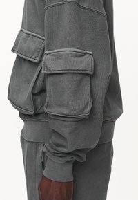 Mörkgrå sweatshirtärm med stor lucka-ficka och ribbad mudd, bärs med matchande byxor på en mörkhyad arm.