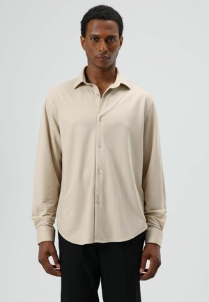 Beige overhemd met lange mouwen van zachte stof, voorzien van een puntkraag, knoopsluiting aan de voorkant en een afgeronde zoom. Gecombineerd met een zwarte broek.