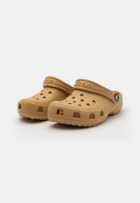 Tan Crocs-kläskor i lätt gummi, med ventilationshål, ett band över insteget och texturerade, halkfria sulor.
