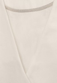 Weißes Oberteil mit einem V-Ausschnitt und einem dezenten silbernen Streifen entlang des Ausschnitts. Strukturierter Stoff mit einer glatten, weichen Oberfläche.