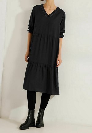 Femme portant une longue robe noire à volants avec des manches retroussées, des leggings noirs et des bottines noires, debout contre un mur clair uni.