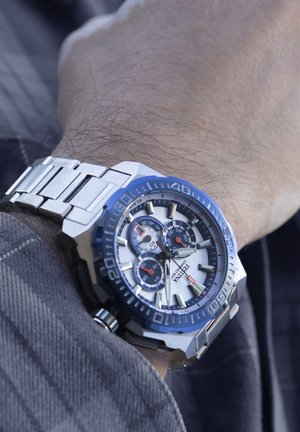 Montre en acier inoxydable avec une lunette bleue, un cadran argenté et trois sous-cadres. Présente des index horaires audacieux et un bracelet à maillons durable.
