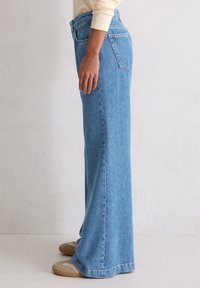 Persoon in wijde blauwe denim jeans en beige sneakers, staand tegen een effen lichte muur.