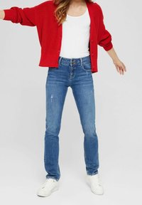 Roter Strickcardigan über einem weißen T-Shirt, kombiniert mit blauen Jeans mit ausgefransten Details und weißen Sneakers.