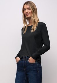 Blonde Frau mit Händen in den Taschen, die ein schwarzes Langarmoberteil und dunkelblaue Jeans trägt, vor einem schlichten hellen Hintergrund stehend.