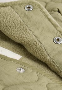 Innenraum einer olivgrünen gesteppte Jacke mit weichem Fleece-Futter, silbernen Druckknöpfen und gerippten Akzenten an den Kanten. Sichtbares Detail des strukturierten Stoffes.