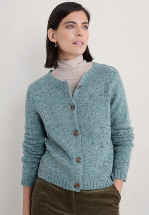 Vrouw met gouden oorbellen, beige coltrui, blauwe cardigan met knopen en bruine corduroy broek, die iets naar rechts kijkt met haar handen in de zakken.