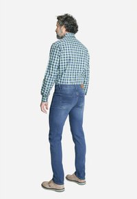 Camisa de cuadros para hombre en tonos verdes y azules, combinada con unos jeans azul ajustados. Zapatos beige con suelas oscuras. Vista trasera.