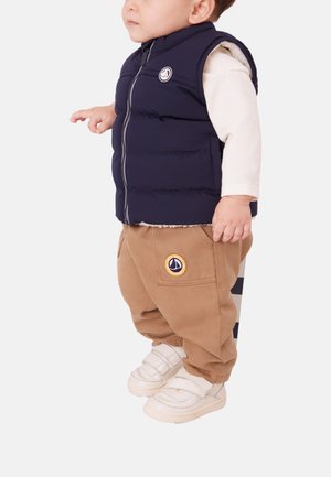 Temno modra puhasta telovnik z zadrgo in logotipom, bela majica z dolgimi rokavi, rjave hlače cargo z emblemom na žepu ter bele čevlje z ježkom.